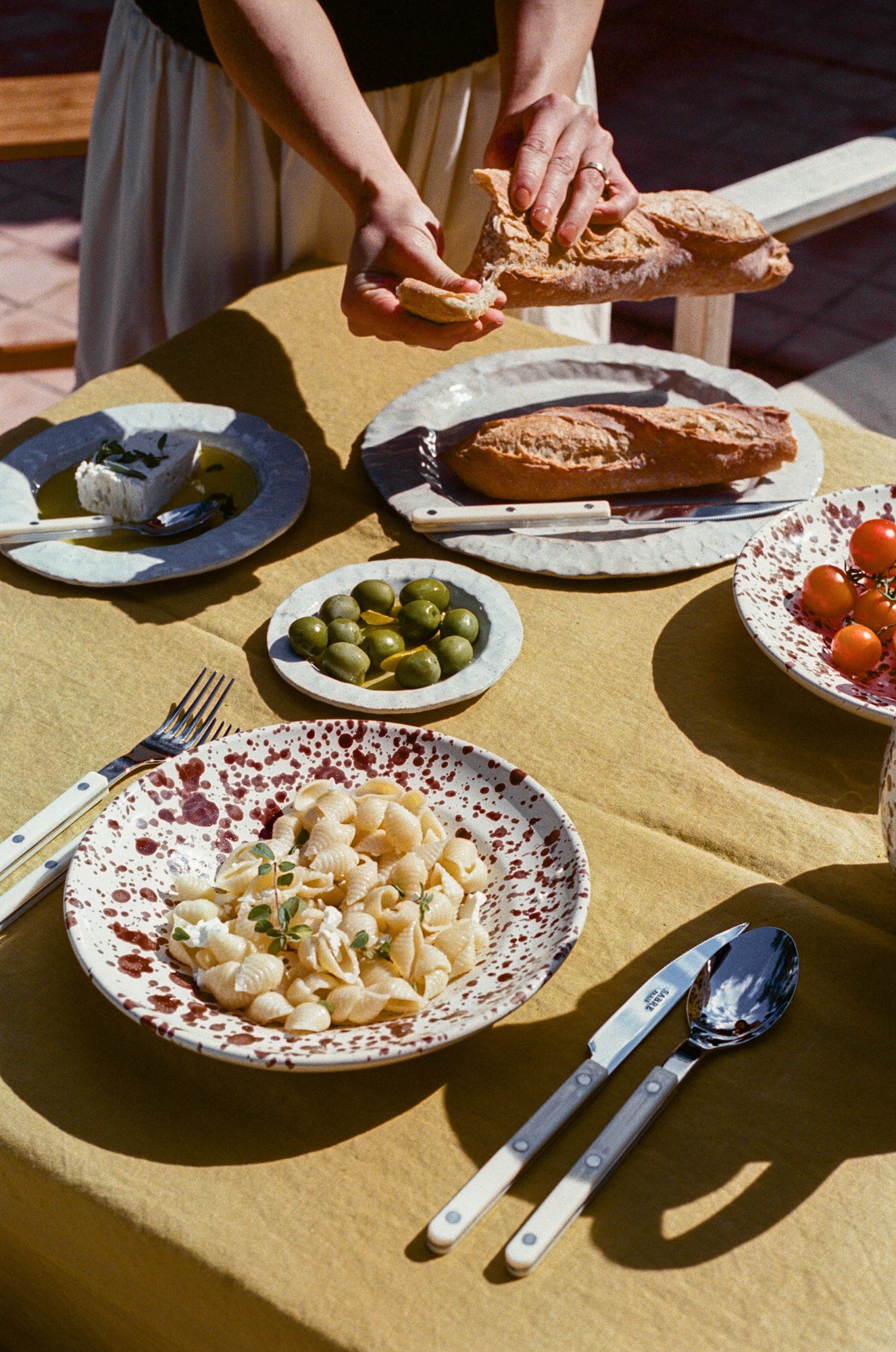Tablescape featuring speckled ceramic deep plate, white ceramic plates, enamel flatware and an assortment of food.