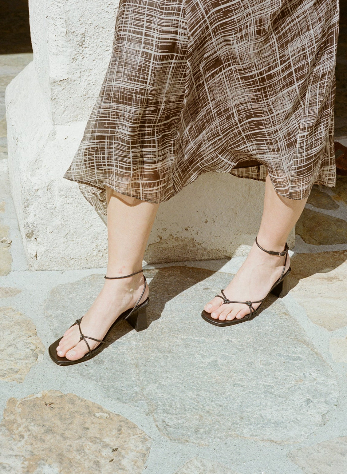 Model wears black high heeled leather sandal with thin knotted straps and ankle wrap buckle closure.