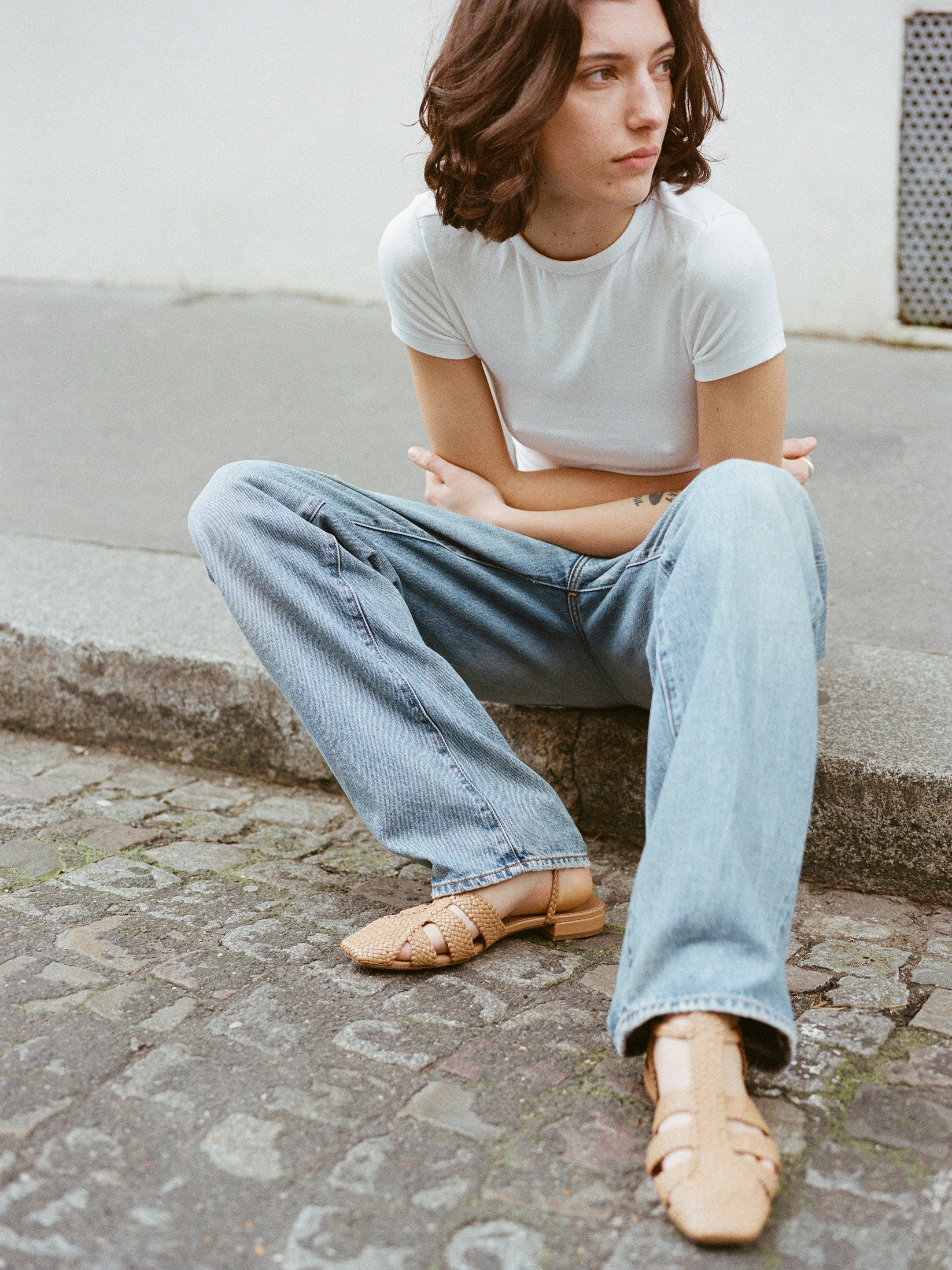 Model wears tan leather fisherman-style sandals featuring straps made of woven leather across the foot and a strap that buckles around the ankle