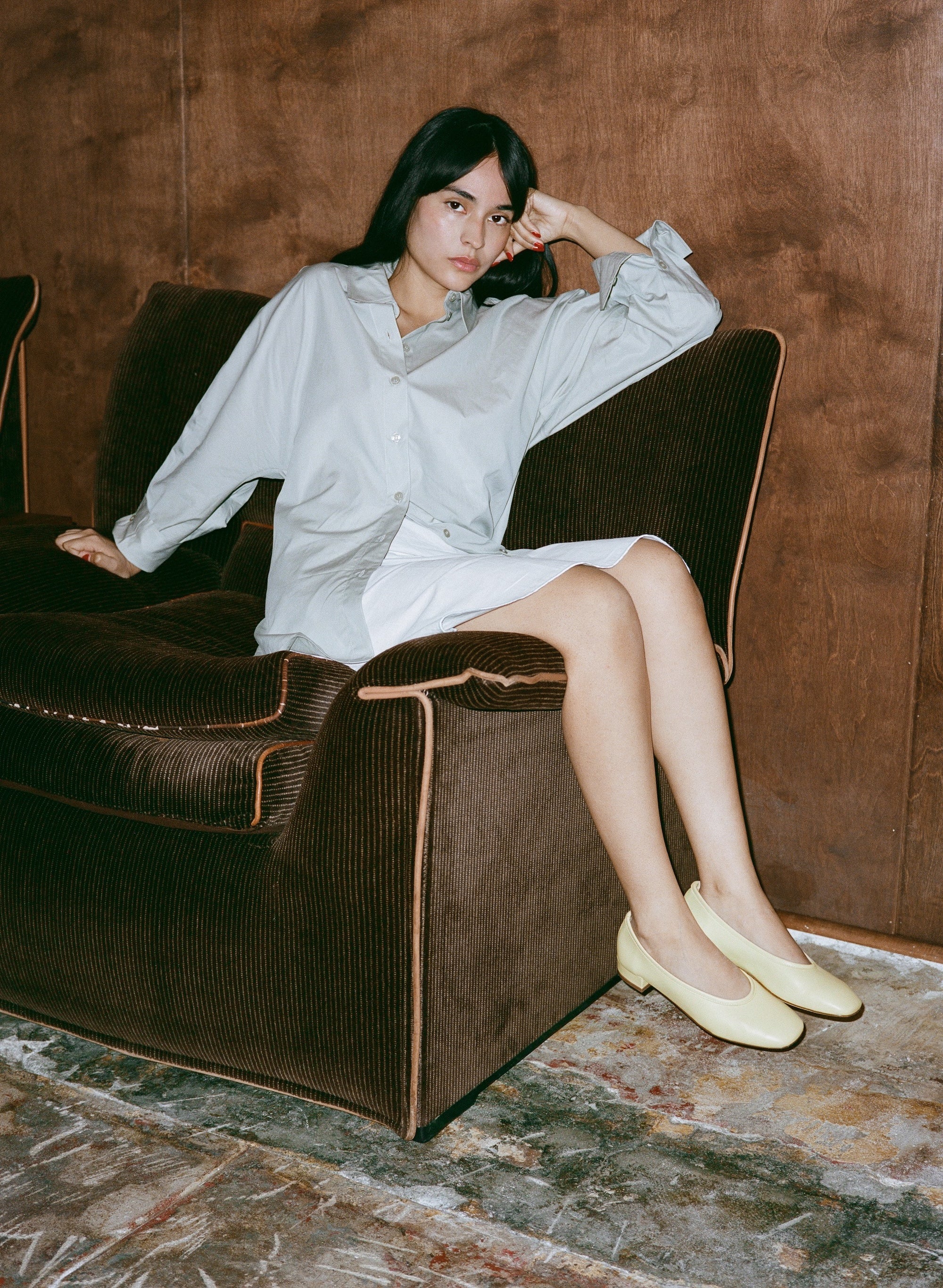 Woman in a light shirt and light yellow pumps, sitting on a brown armchair with a wooden wall background