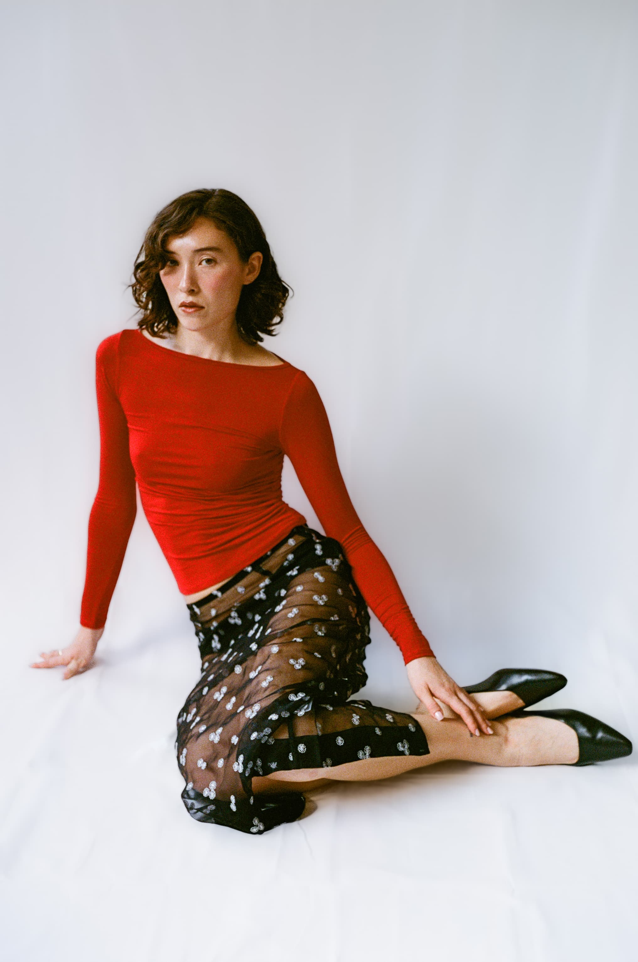Woman wearing a red top and black mules sitting on a white background