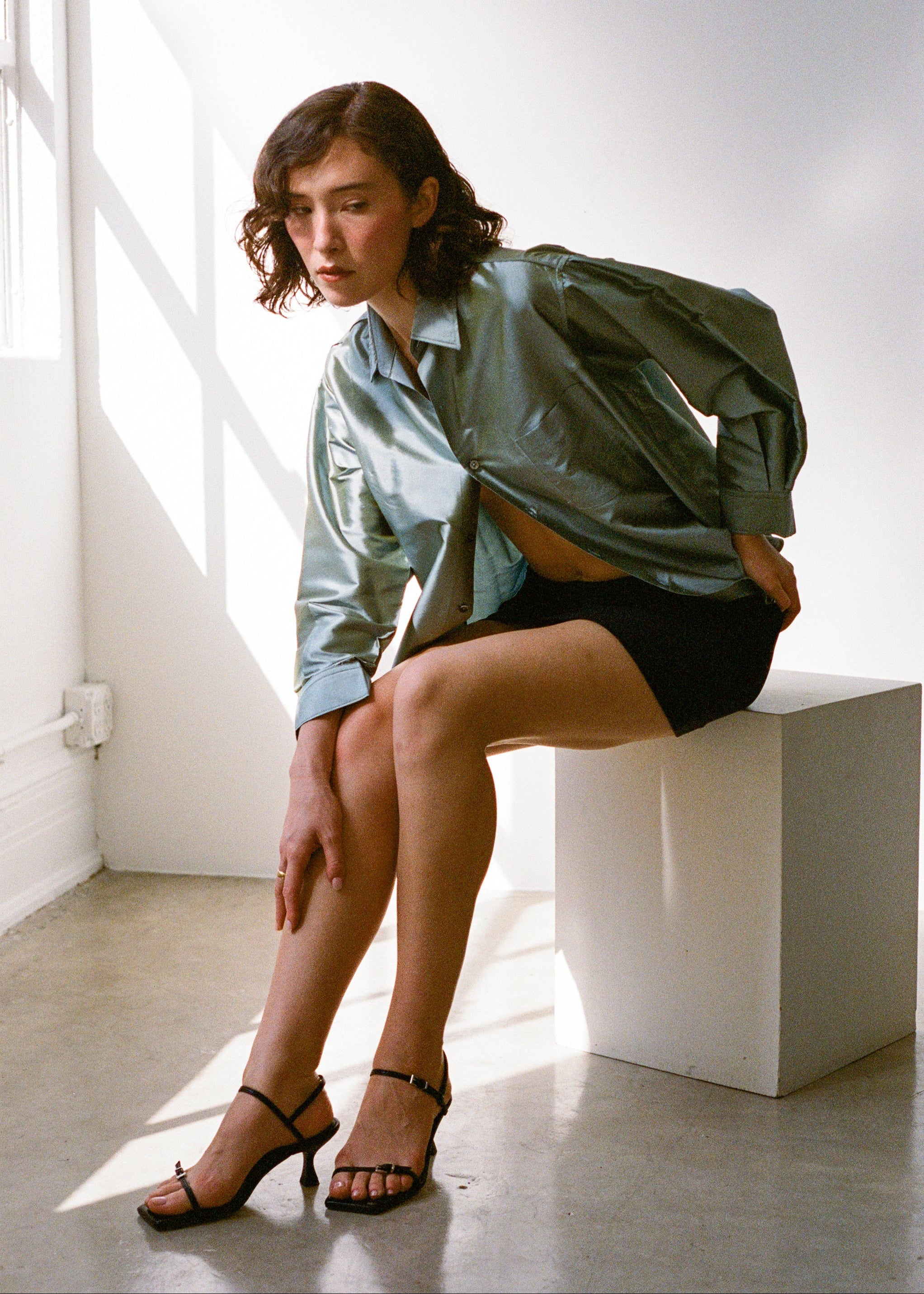 Woman sitting on a white block in a bright room wearing a light blue shirt and black sandals