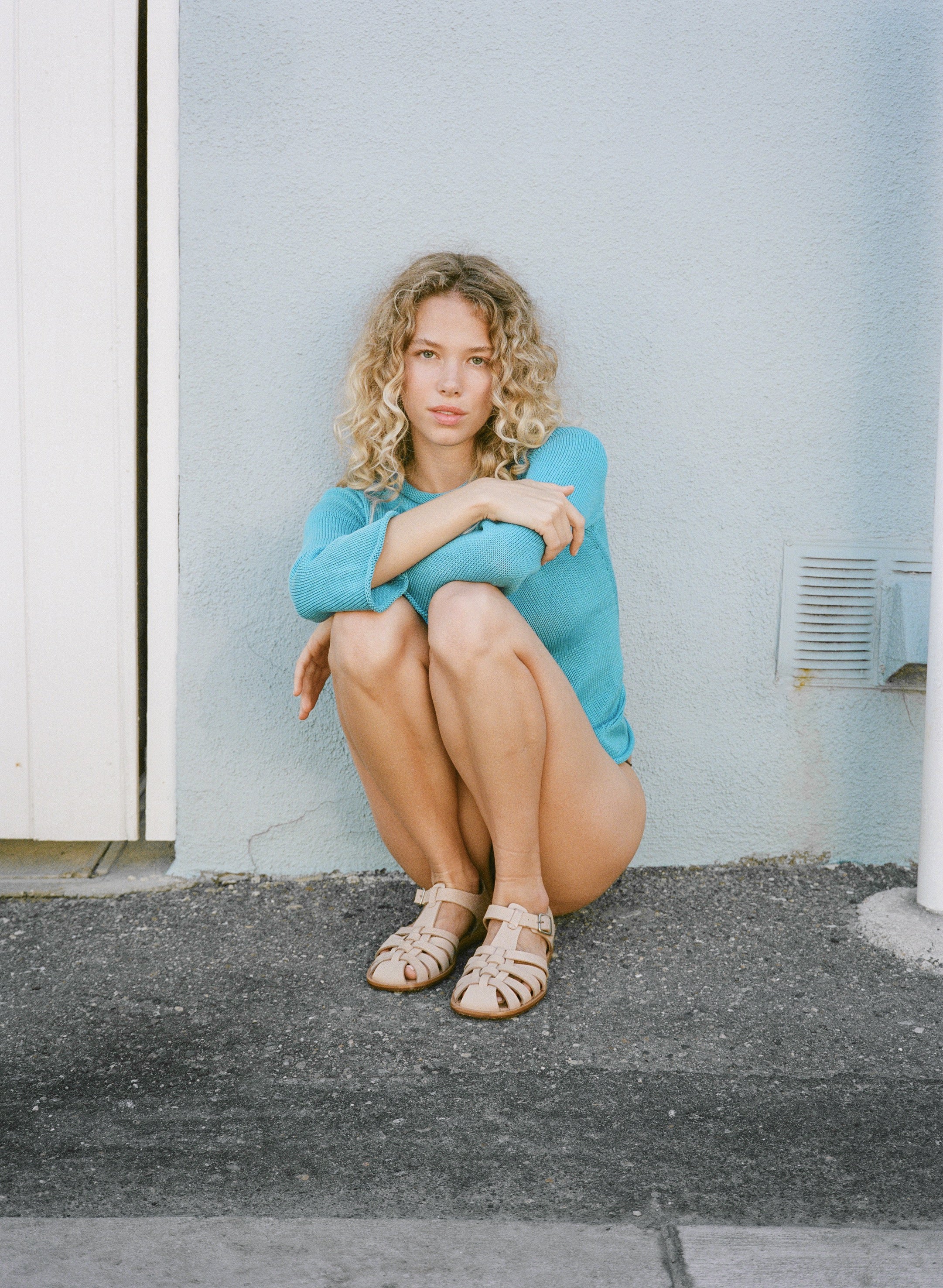 Woman in a blue sweater sitting against a light gray wall.