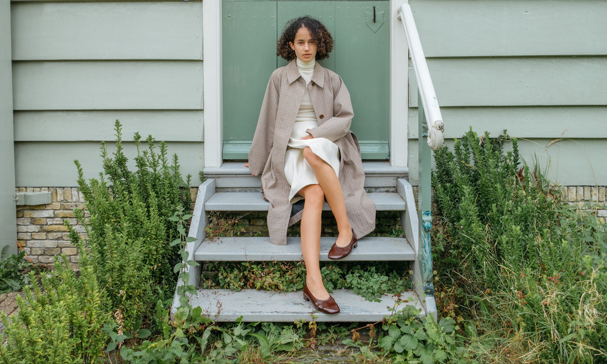 A model sits on the wooden steps of a house wearing a light beige trench coat over a white turtleneck and skirt, with brown leather shoes. The house has pale green siding and a green door, surrounded by green plants.