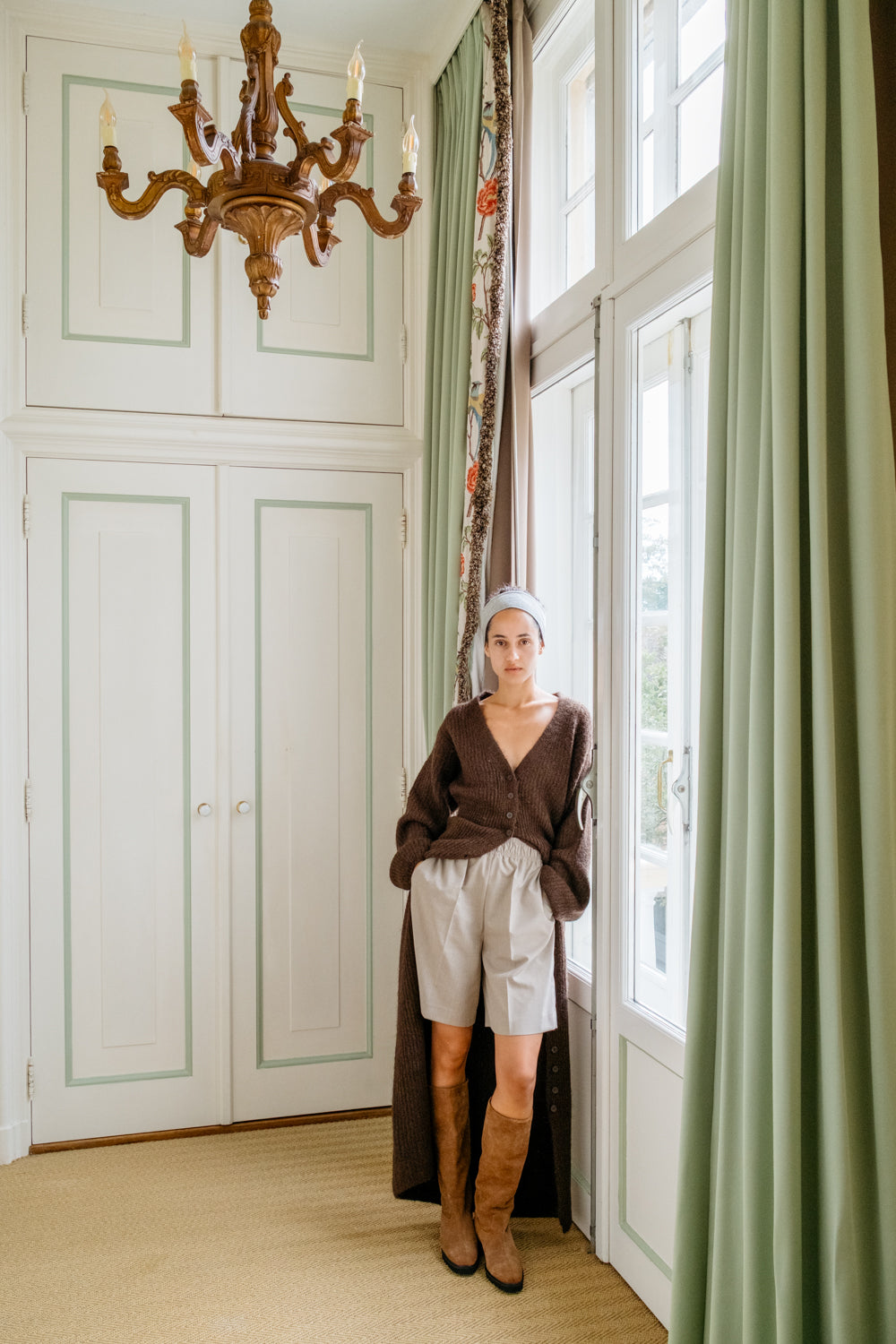 A woman stands in a bright room with tall windows, green curtains, and a chandelier, wearing a brown cardigan, light gray shorts, and brown knee-high boots.