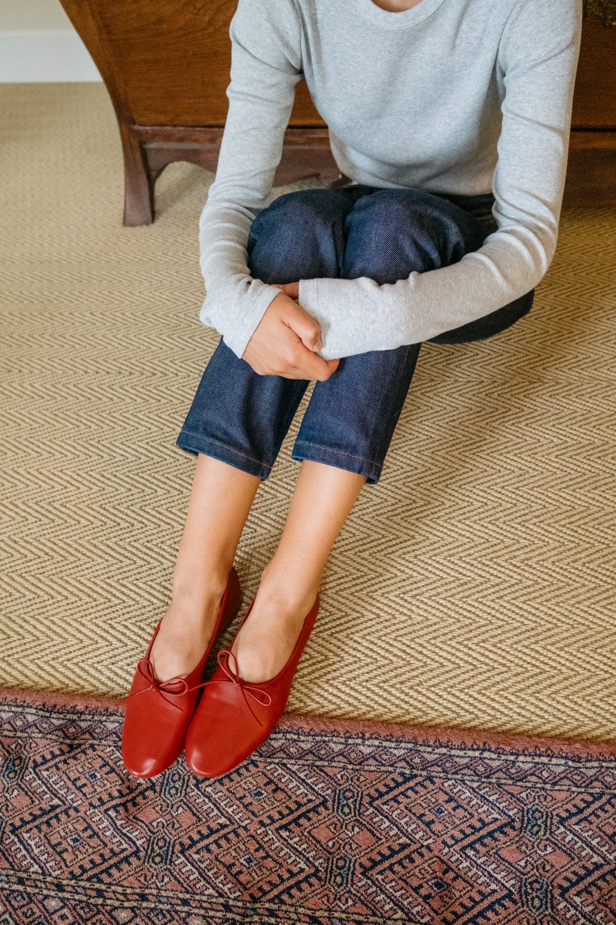 A person sitting on a woven beige rug wearing dark blue jeans, a light gray long-sleeve top, and red leather lace-up flats, with a patterned rug and wooden furniture visible.