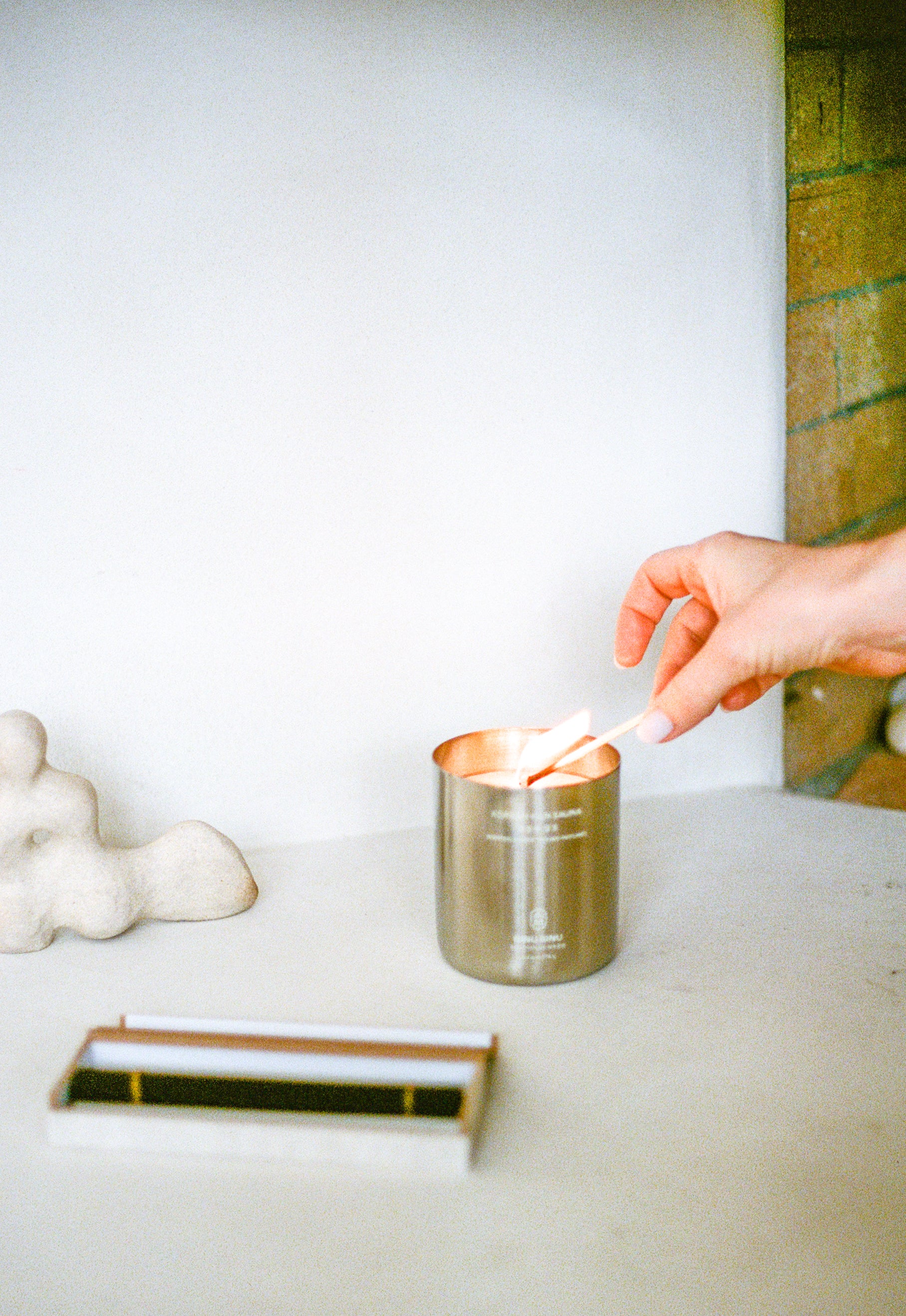 Candle in metal container being lit by model on surface with incense and sculpture.