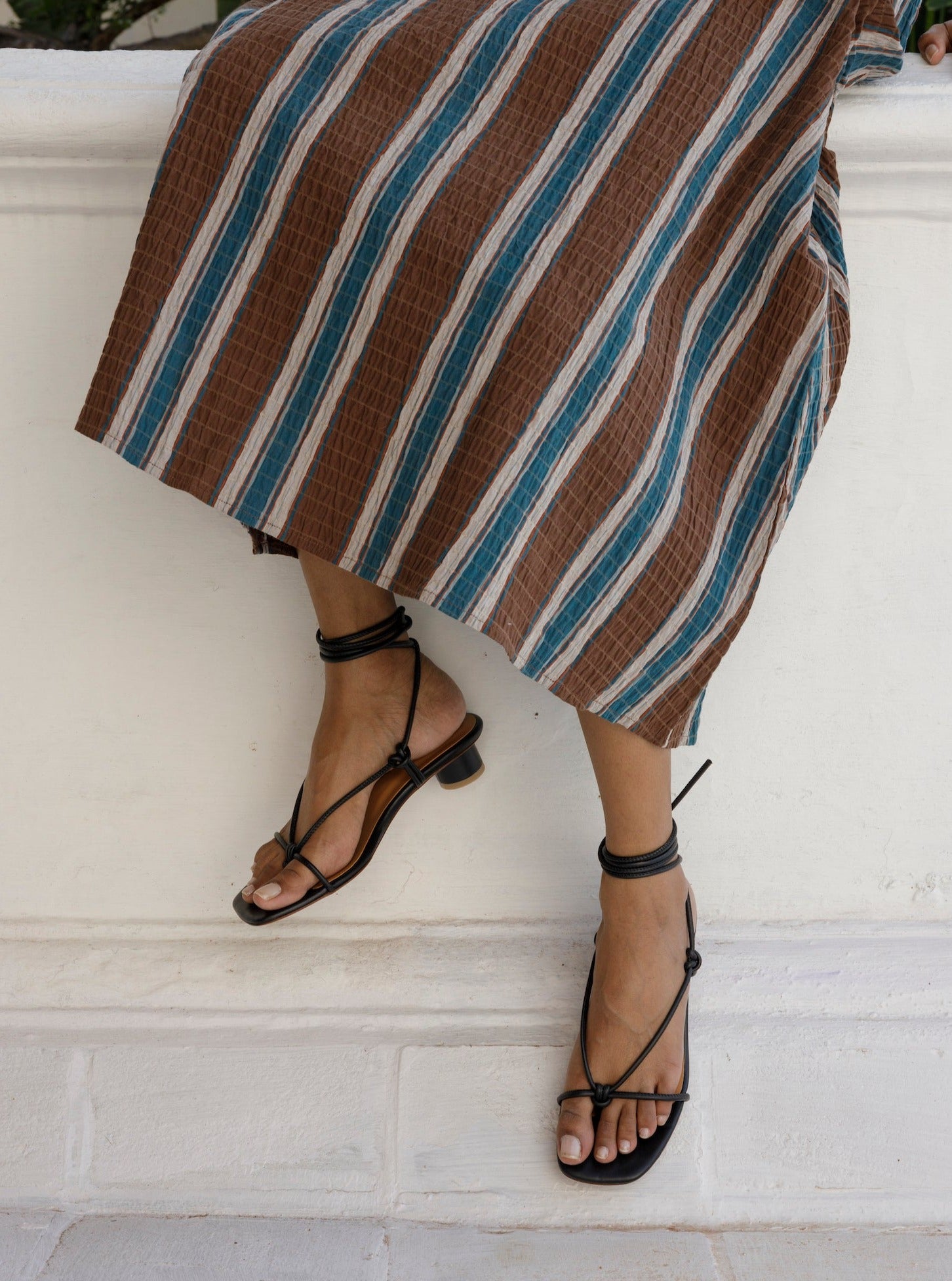 Knotted thong sandal featuring a short heel and tubular black leather straps that wrap around the ankle to tie.