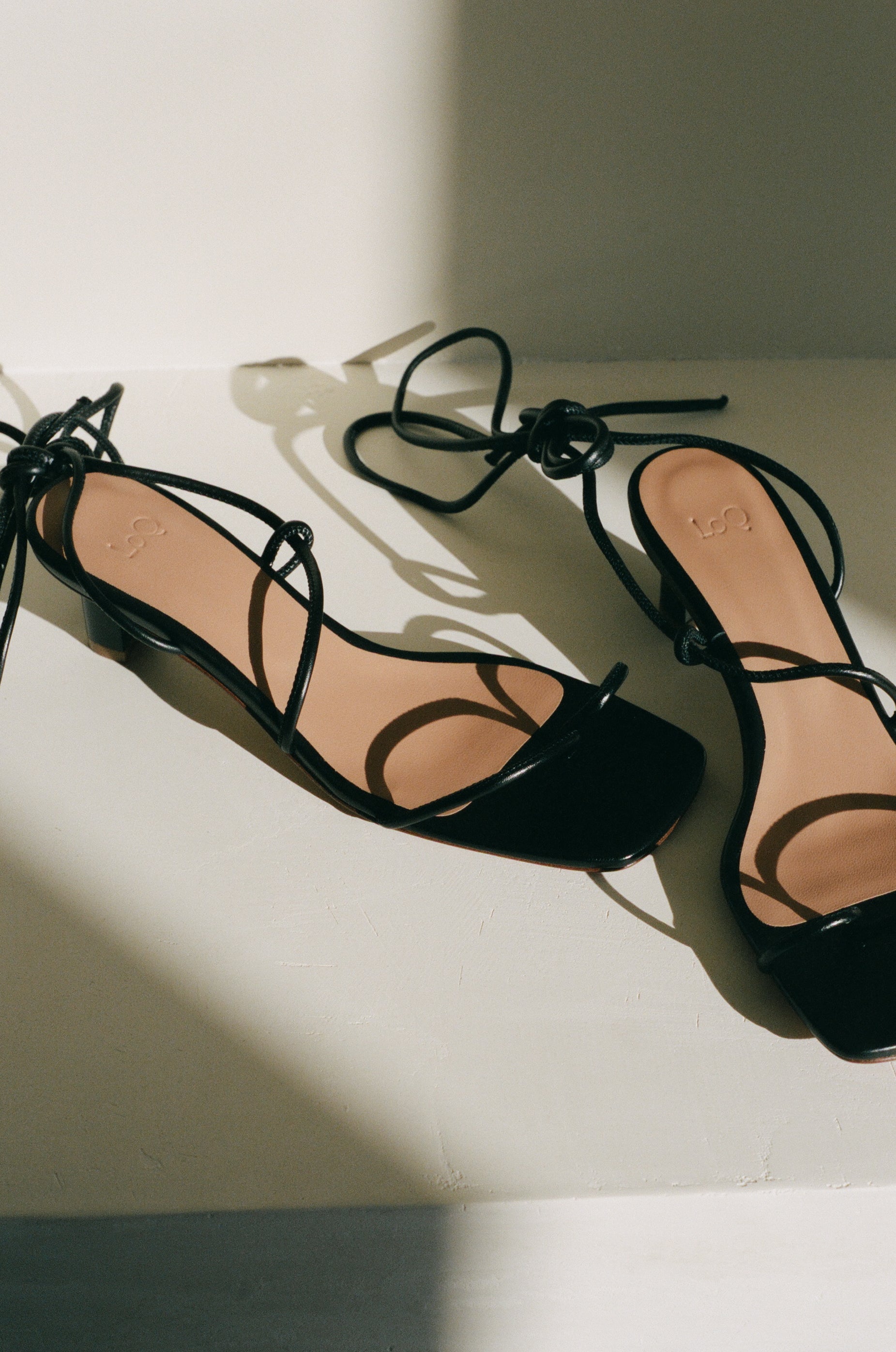 Heeled sandal in black leather featuring tubular leather straps that tie at the ankle and a small sculptural 2" heel; a minimal strap divides at the toe.