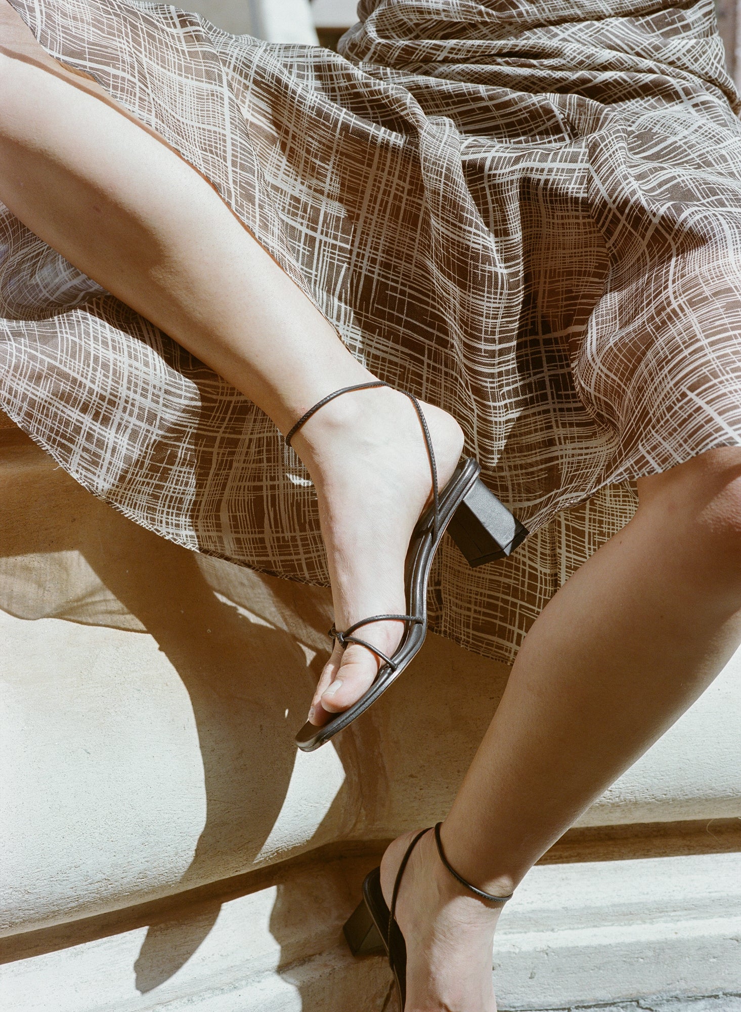 Model wears black high heeled leather sandal with thin knotted straps and ankle wrap buckle closure.