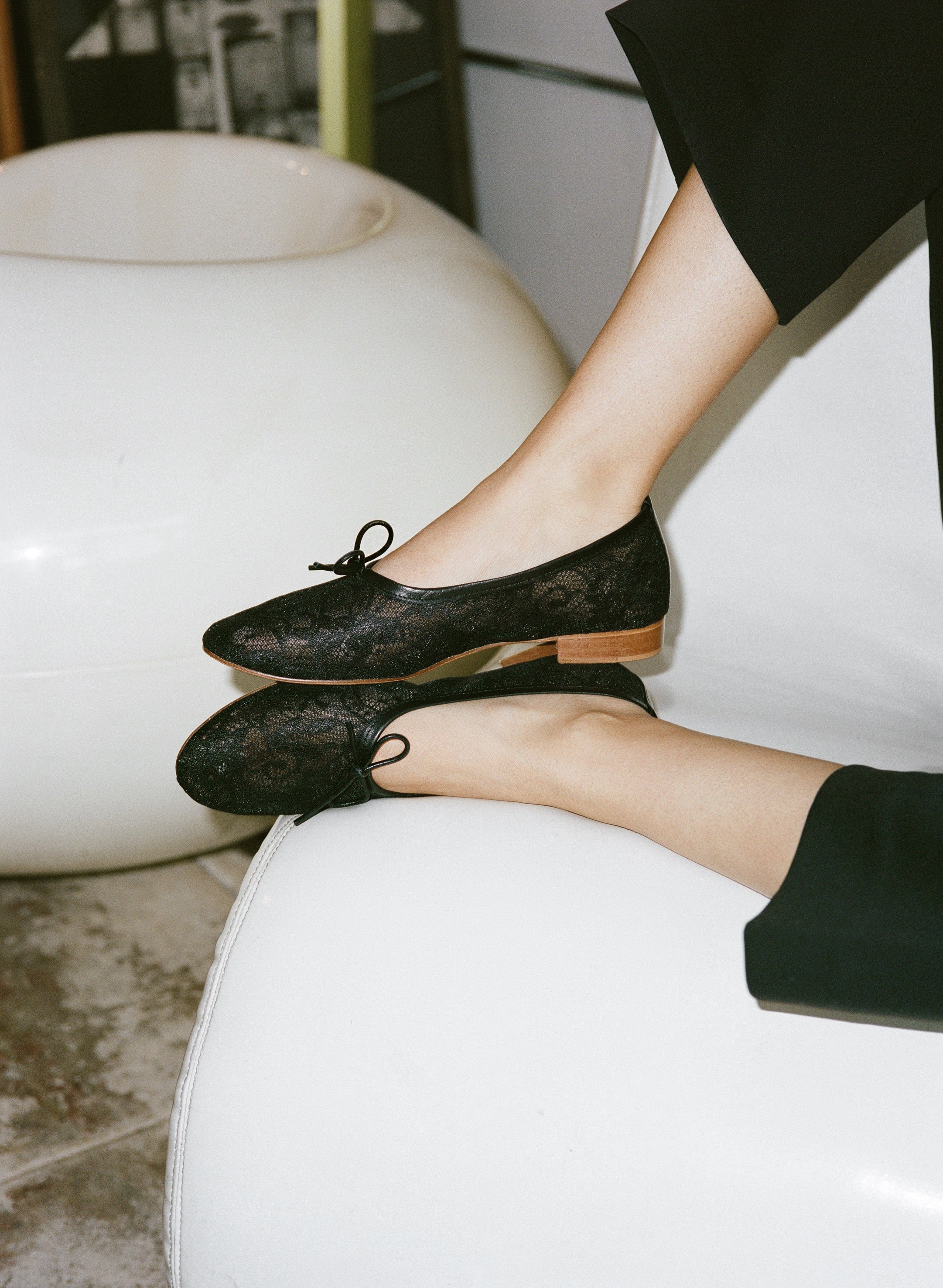 Black lace ballet flats worn by a person sitting on a white chair.