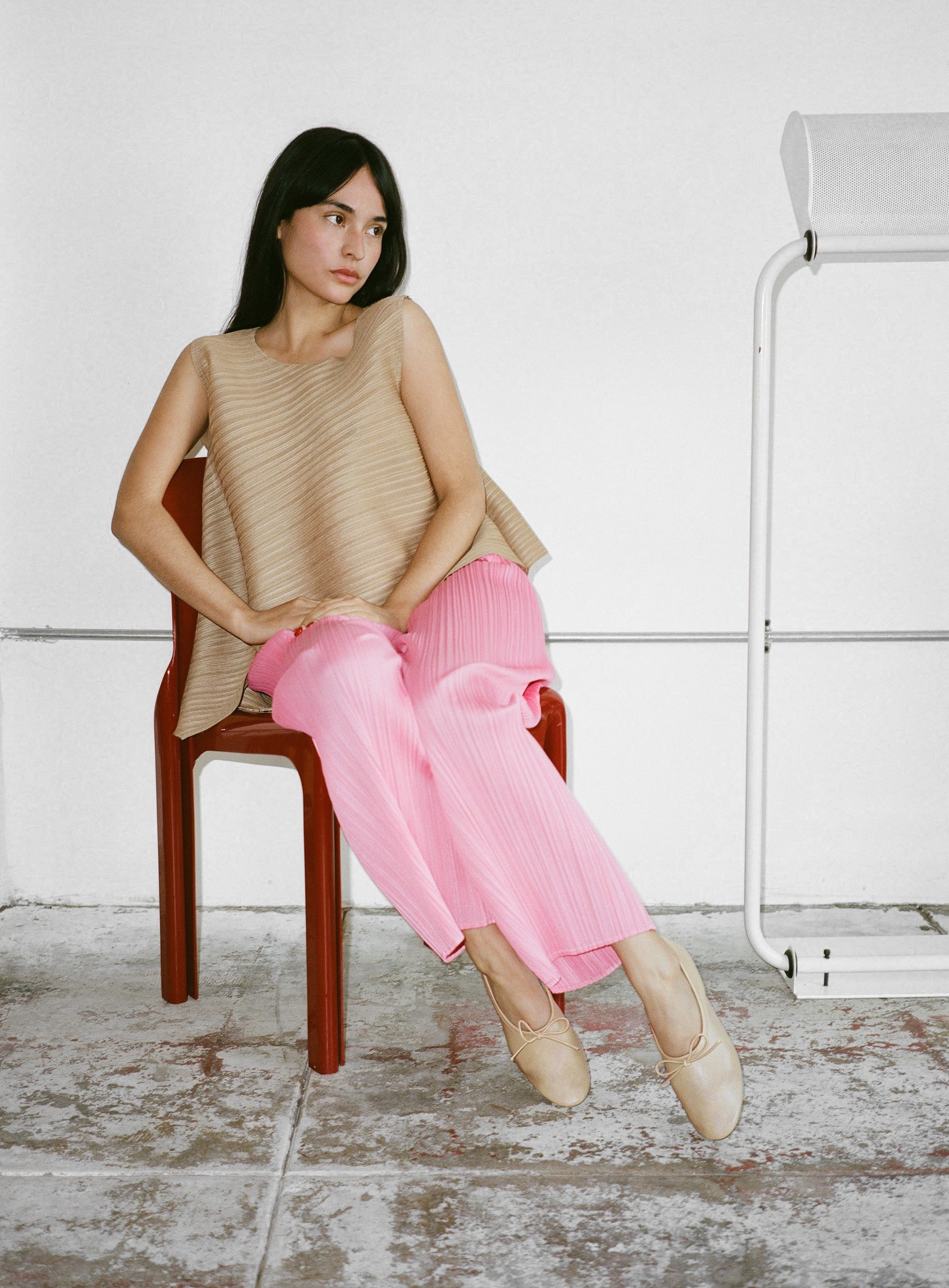 Woman sitting on a chair wearing beige ballet flats and pink pants in a minimalistic setting.