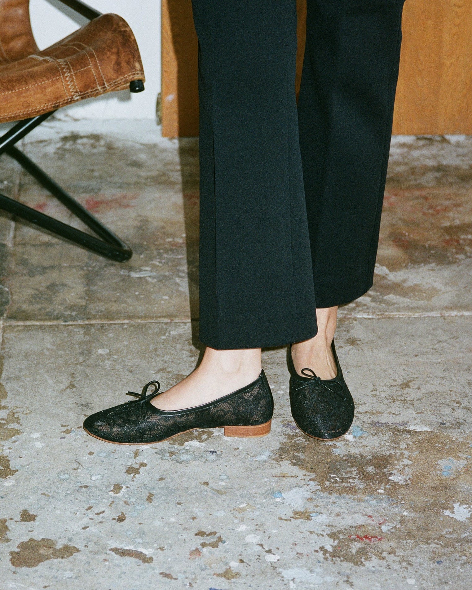 Person wearing black lace flats on a textured floor with a brown leather chair in the background.