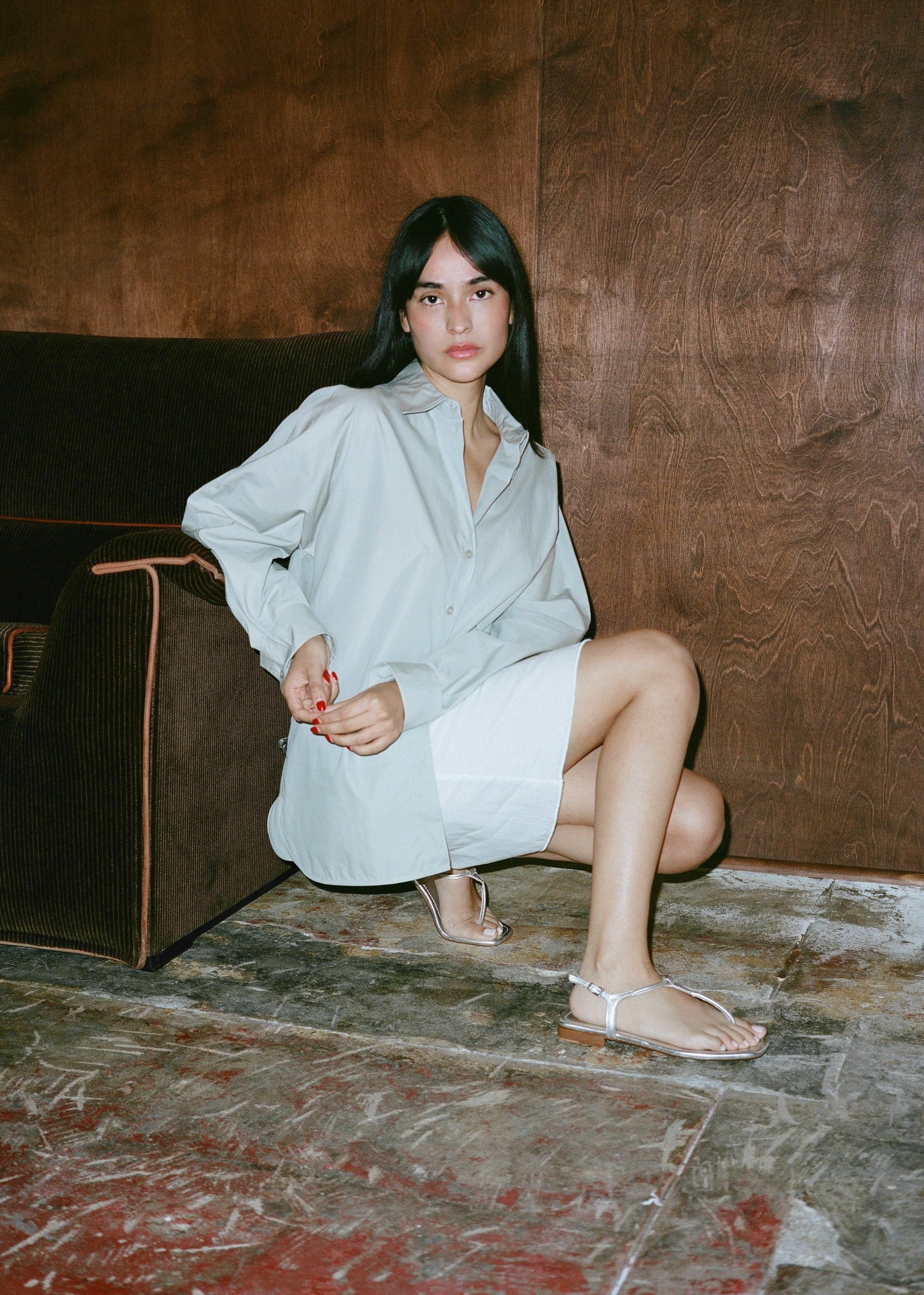 Woman in a light blue shirt and silver sandals on a stone floor against a wooden wall.