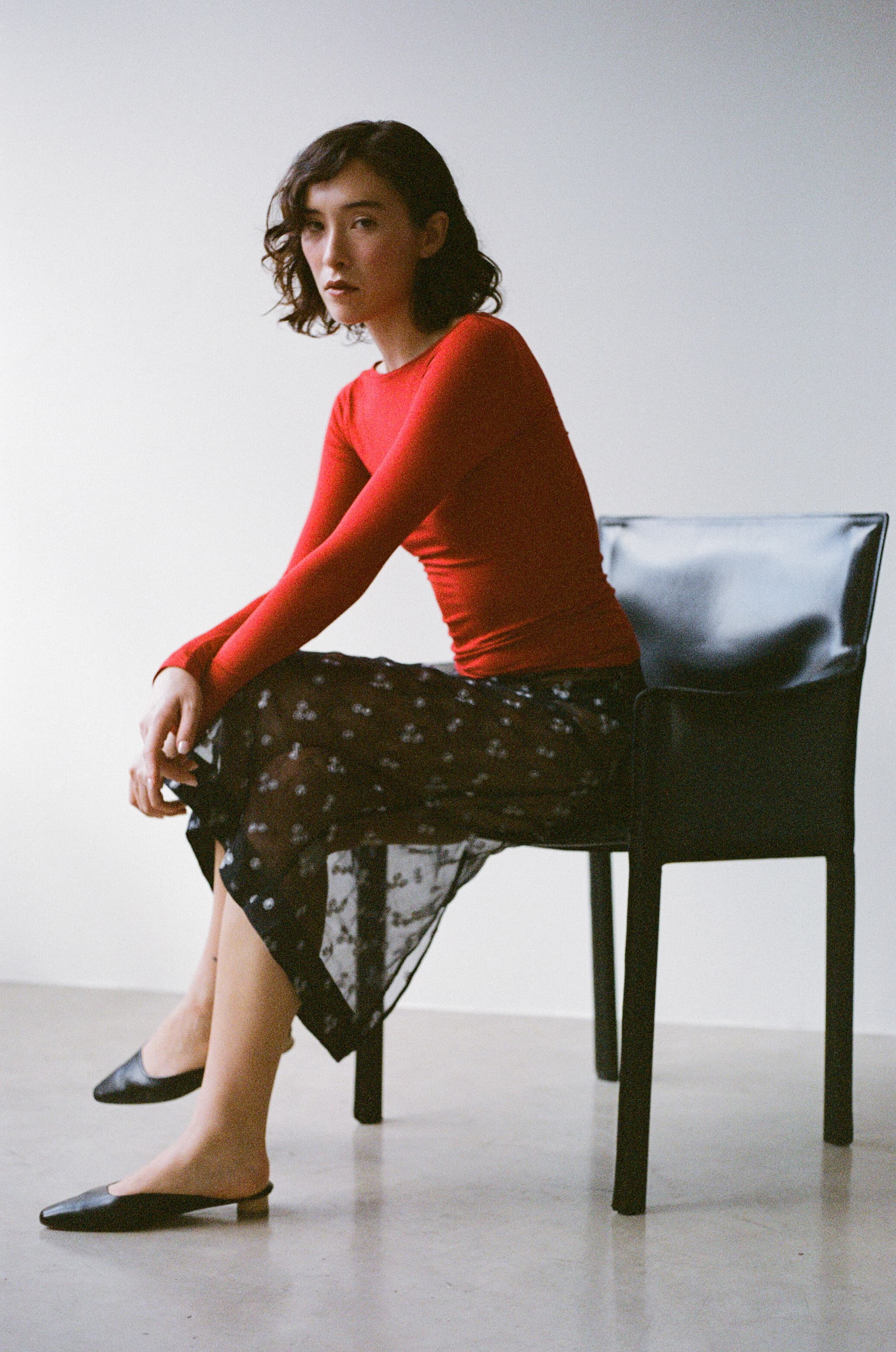 Woman in a red top and black mules on a chair against a white background