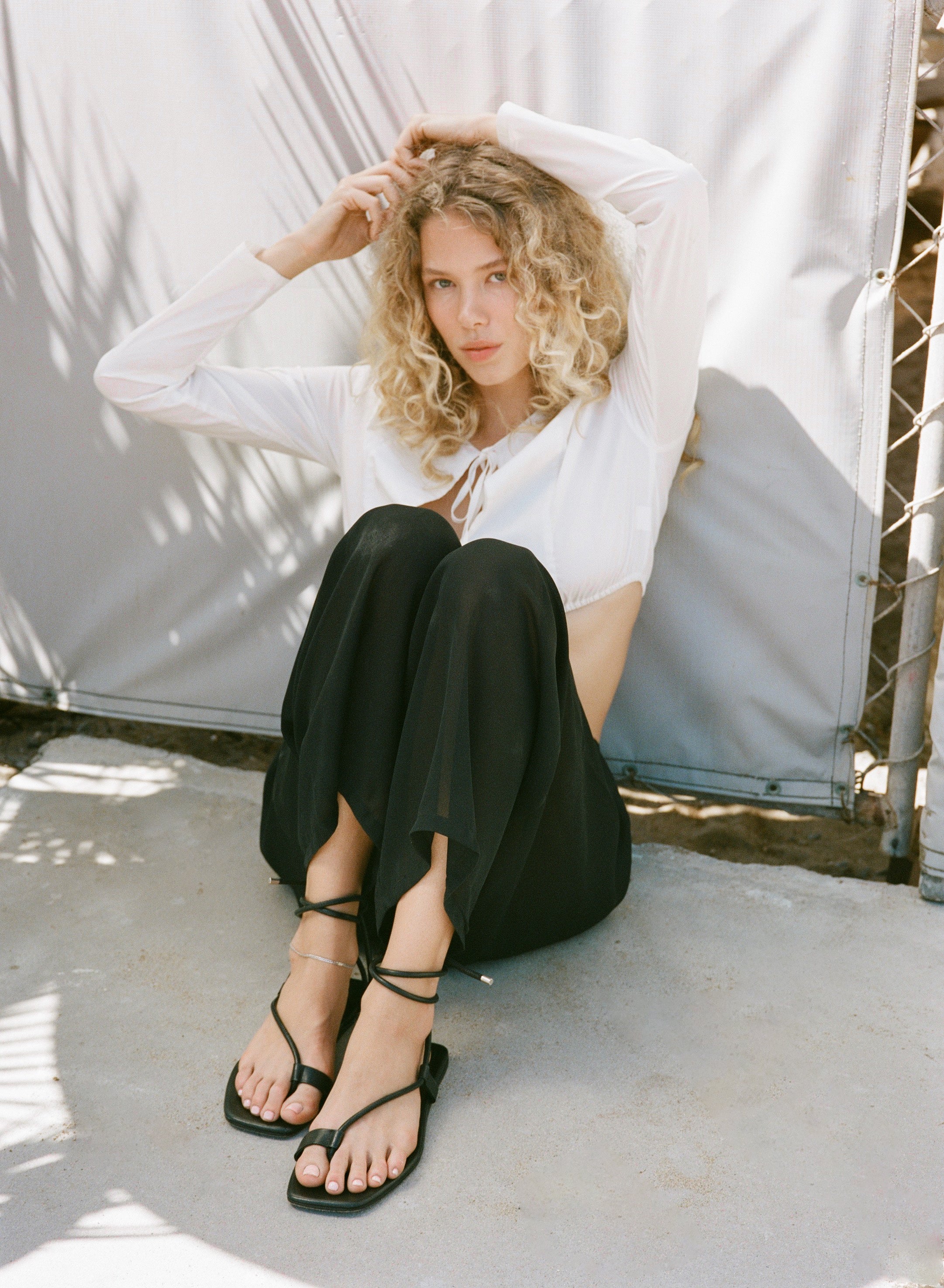 Woman sitting on a concrete surface wearing a white top and black sandals.