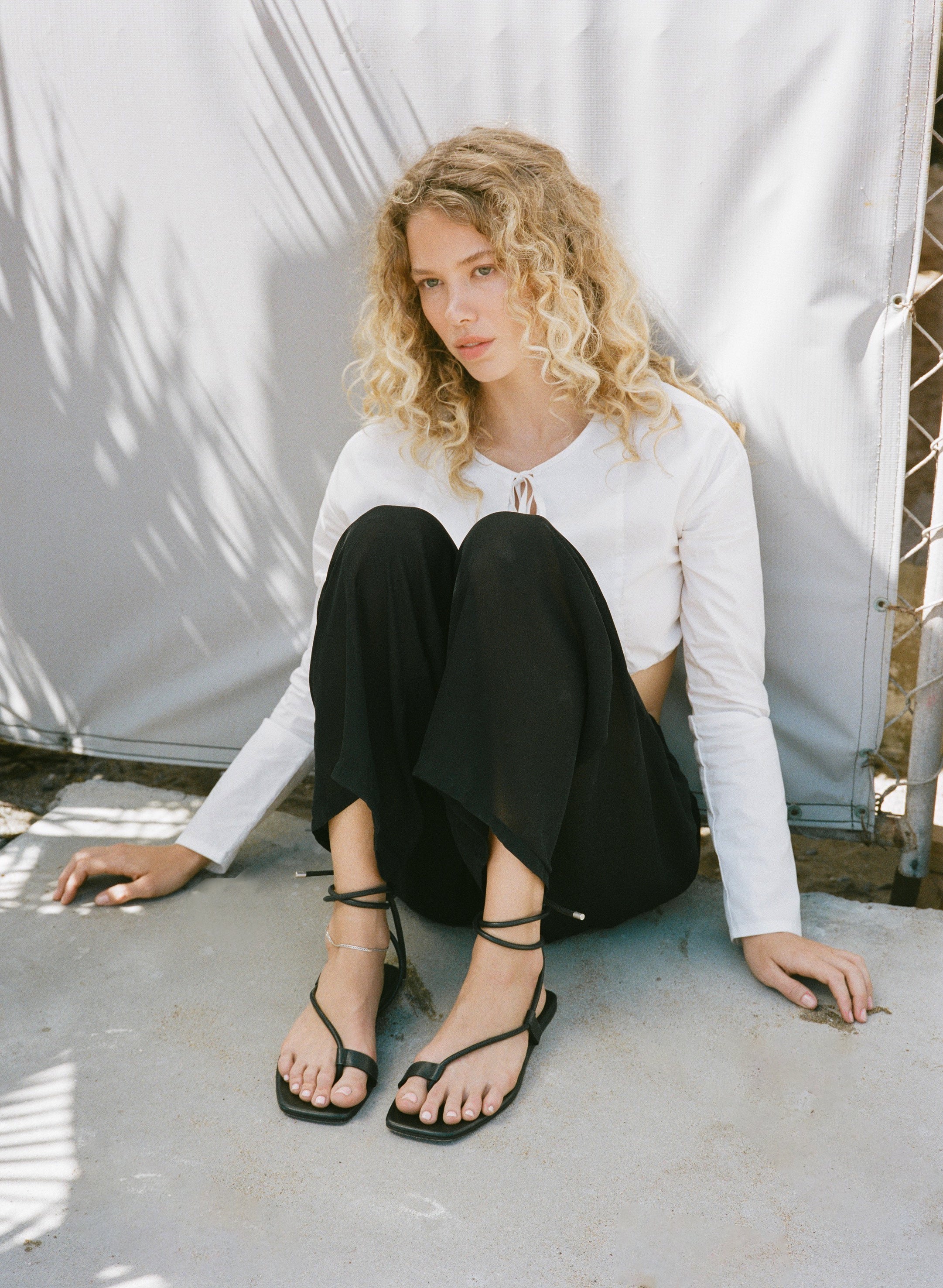 Woman sitting on the ground wearing a white long-sleeve top and black pants with strappy sandals.