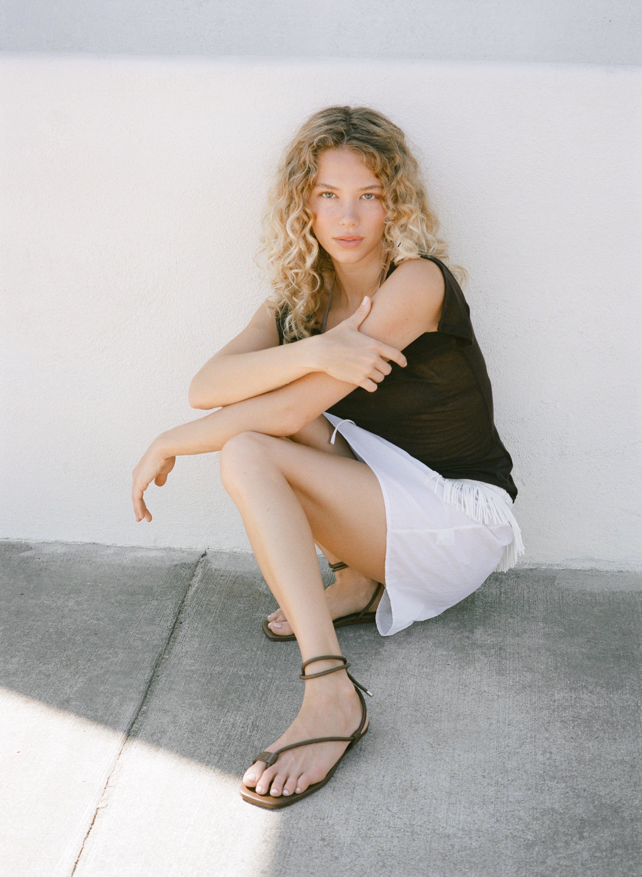 Woman sitting on a concrete floor against a white wall wearing brown strappy sandals