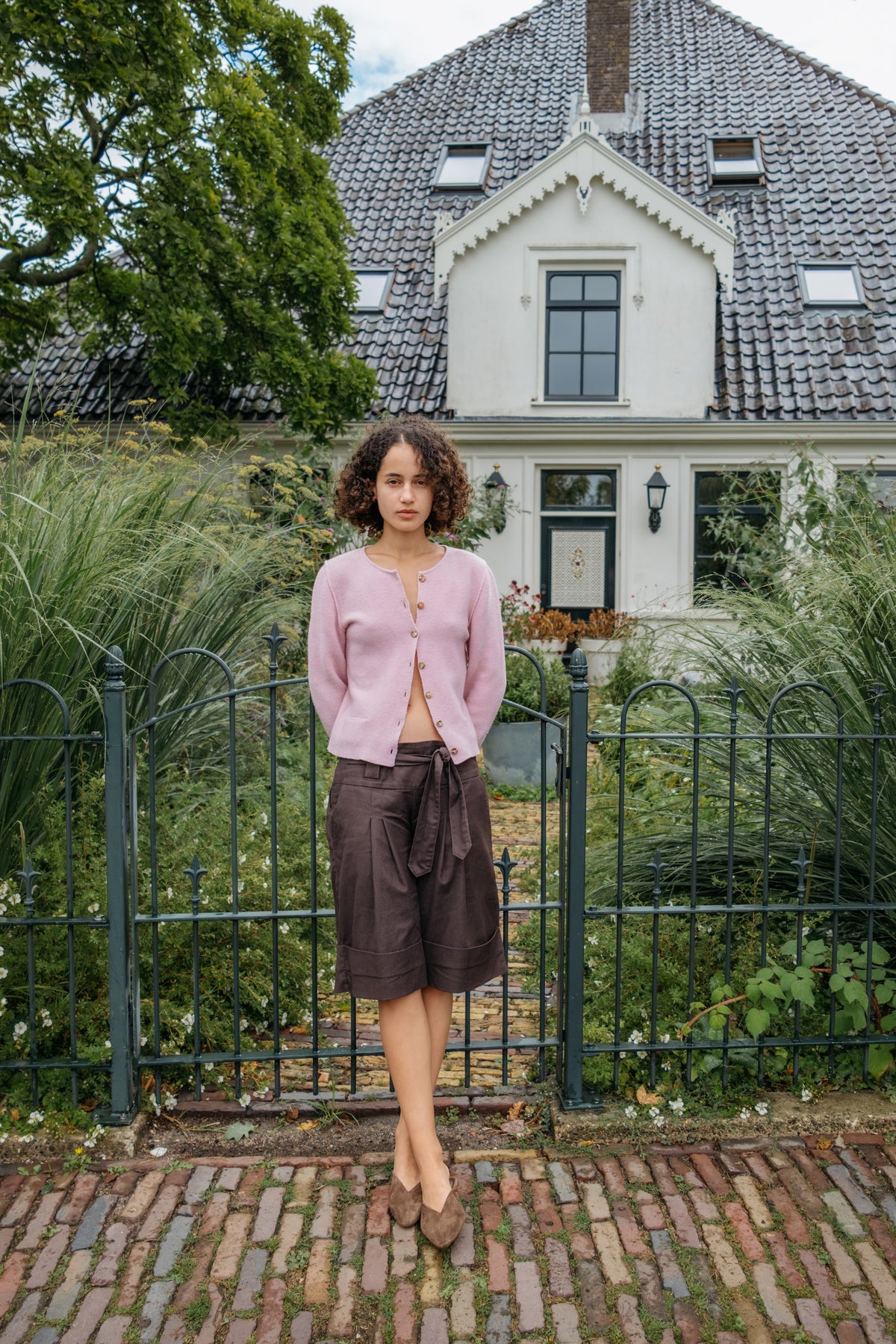 A woman stands outdoors in front of a white house and green fence, wearing a light pink cropped cardigan, dark brown shorts and brown pointed mules.