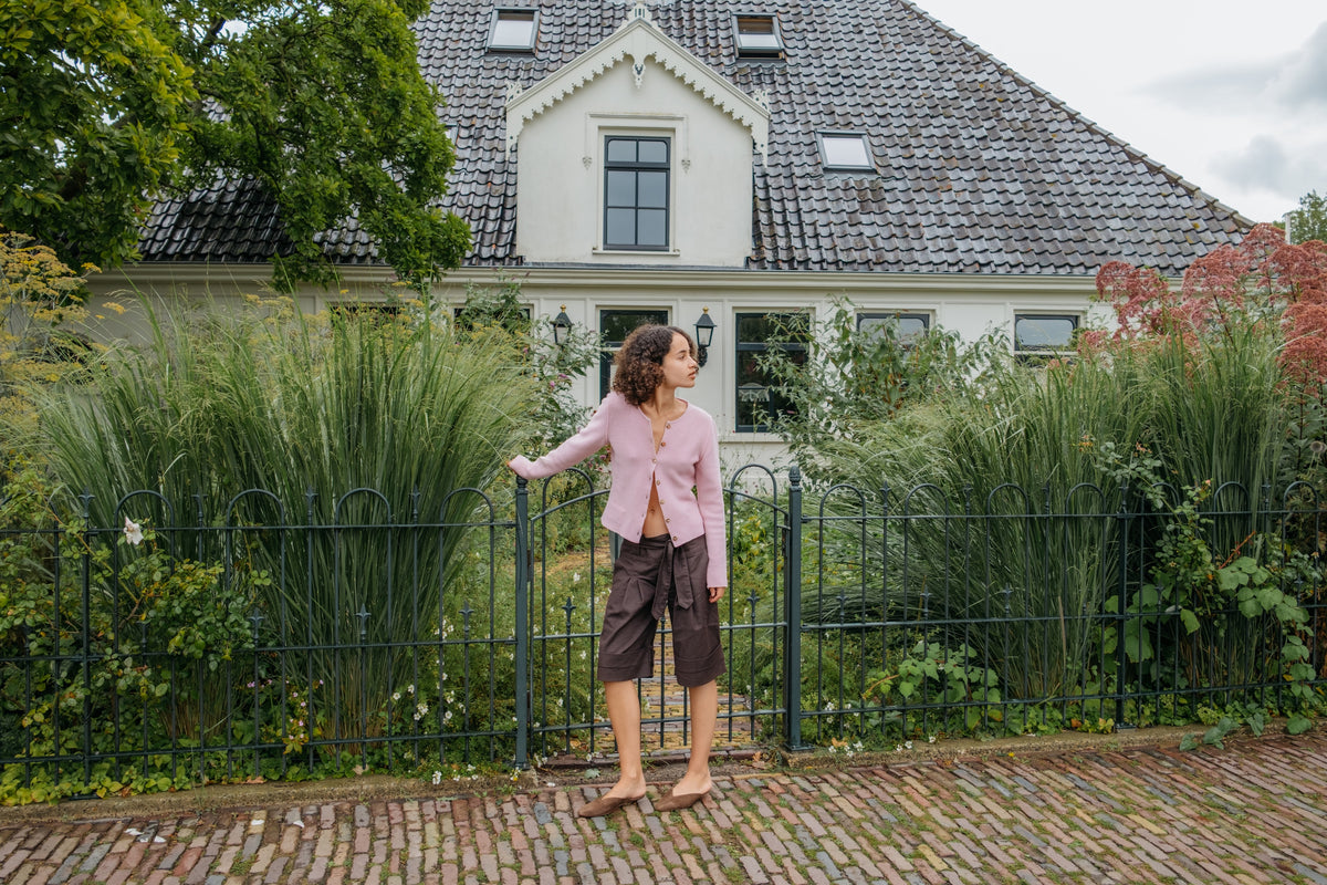 A woman stands barefoot on a cobblestone path in front of a large white house with a dark tiled roof, surrounded by tall ornamental grasses and greenery. She is wearing a light pink cropped cardigan and dark brown suede mules