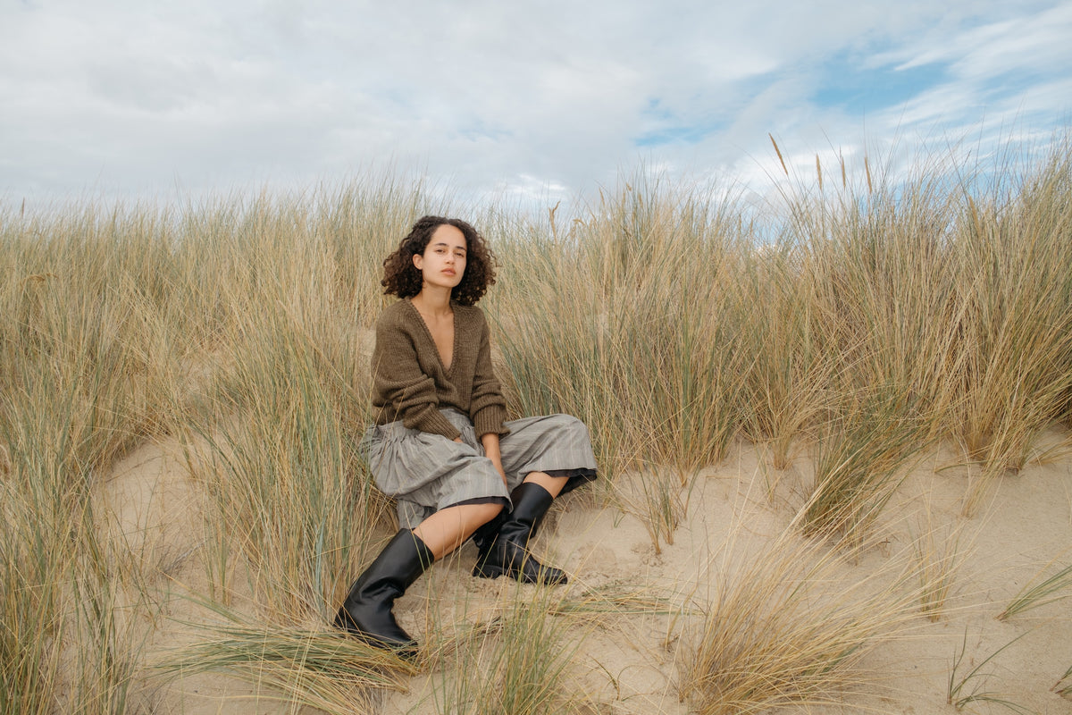 A woman sits on a sandy dune surrounded by tall beach grass under a cloudy sky, wearing black mid calf-high boots.