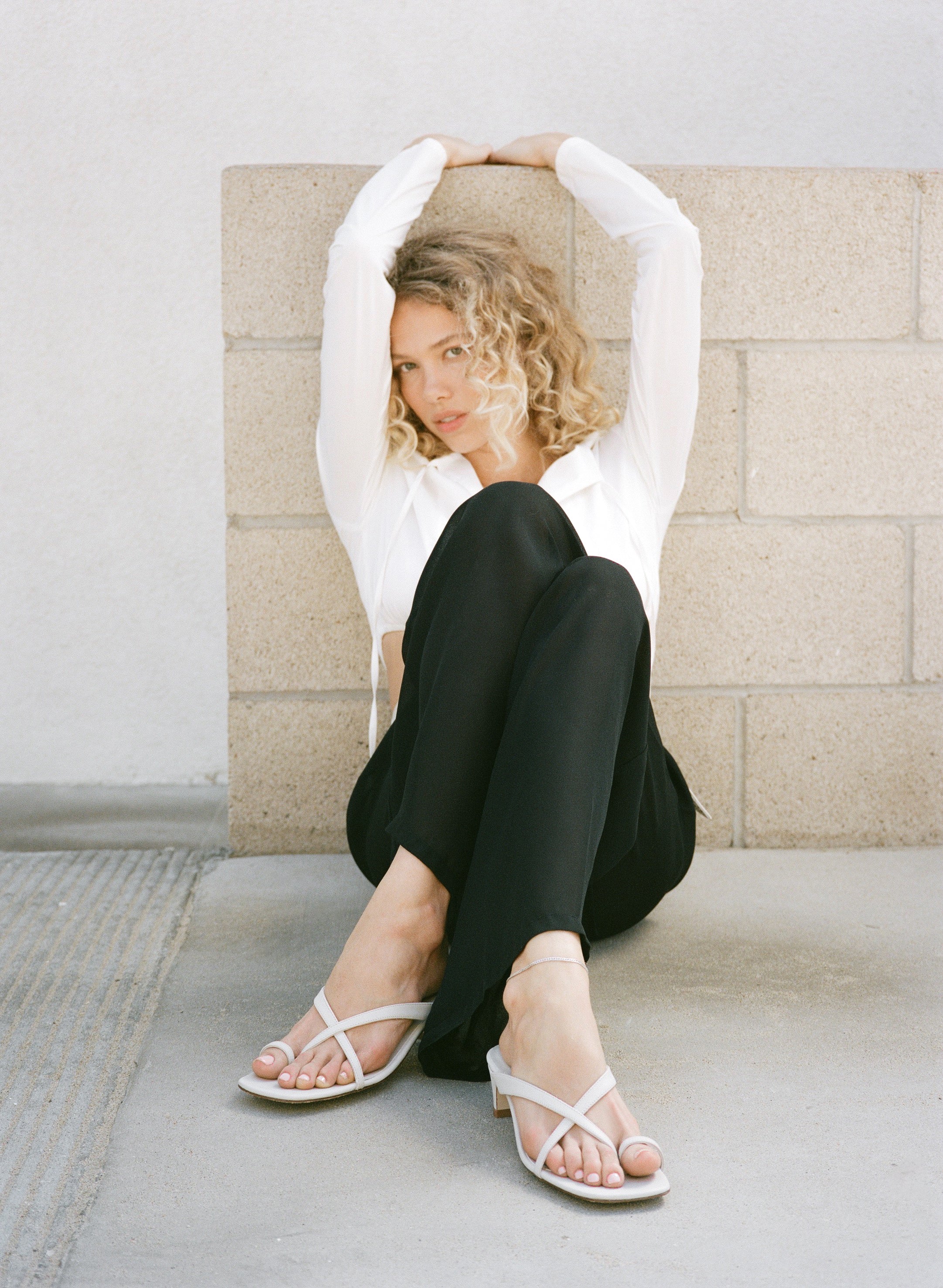 Woman sitting on a stone bench wearing a white shirt, black pants, and white sandals.