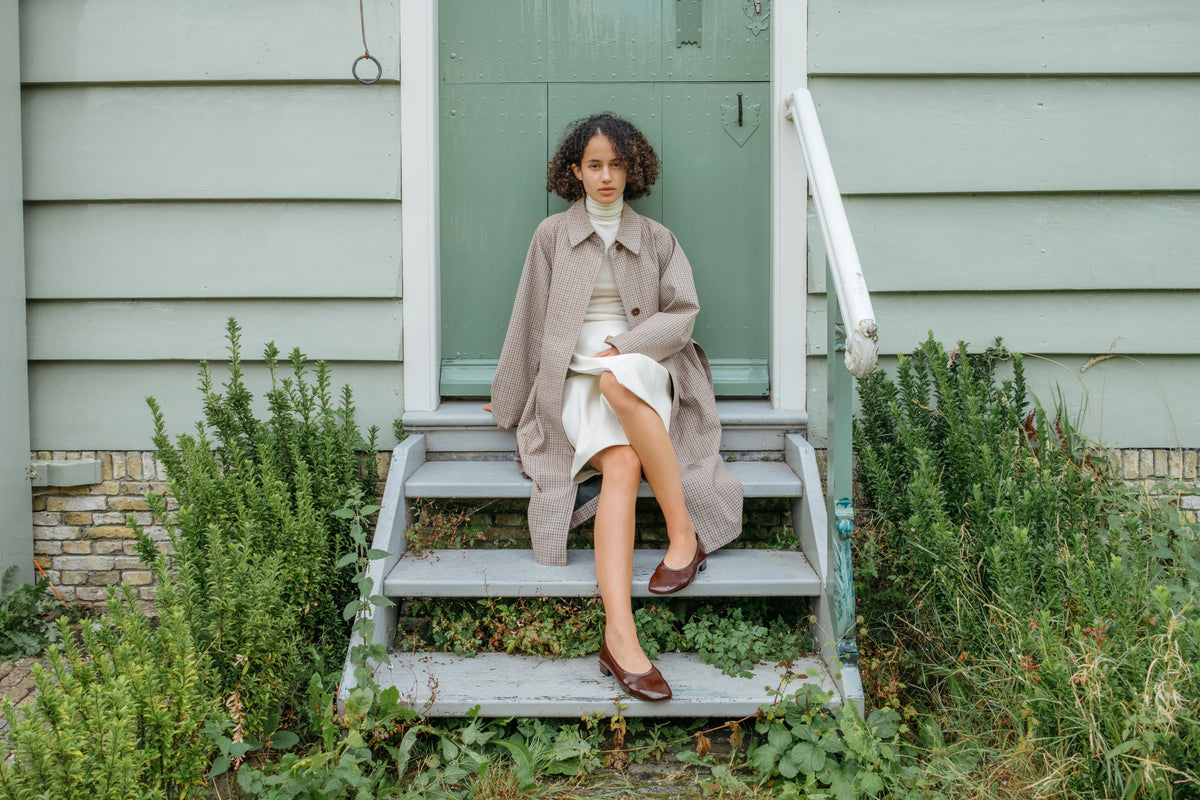 A model sits on the wooden steps of a house wearing a light beige trench coat over a white turtleneck and skirt, with brown leather shoes. The house has pale green siding and a green door, surrounded by green plants.