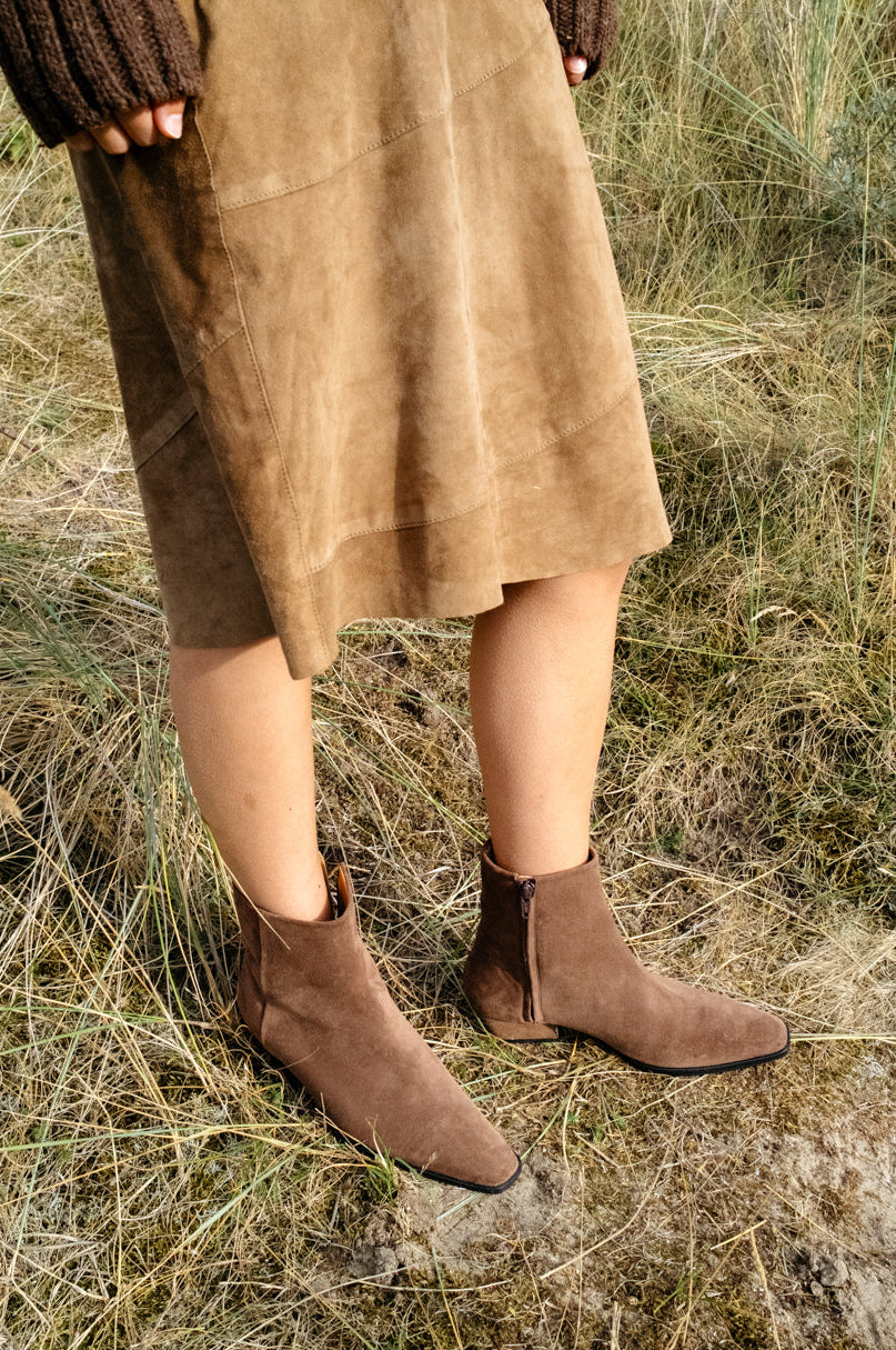 A person standing outdoors in dry grass wearing a pair of coffee brown suede leather western style ankle boots with pointed toes and low cuban heel. 
