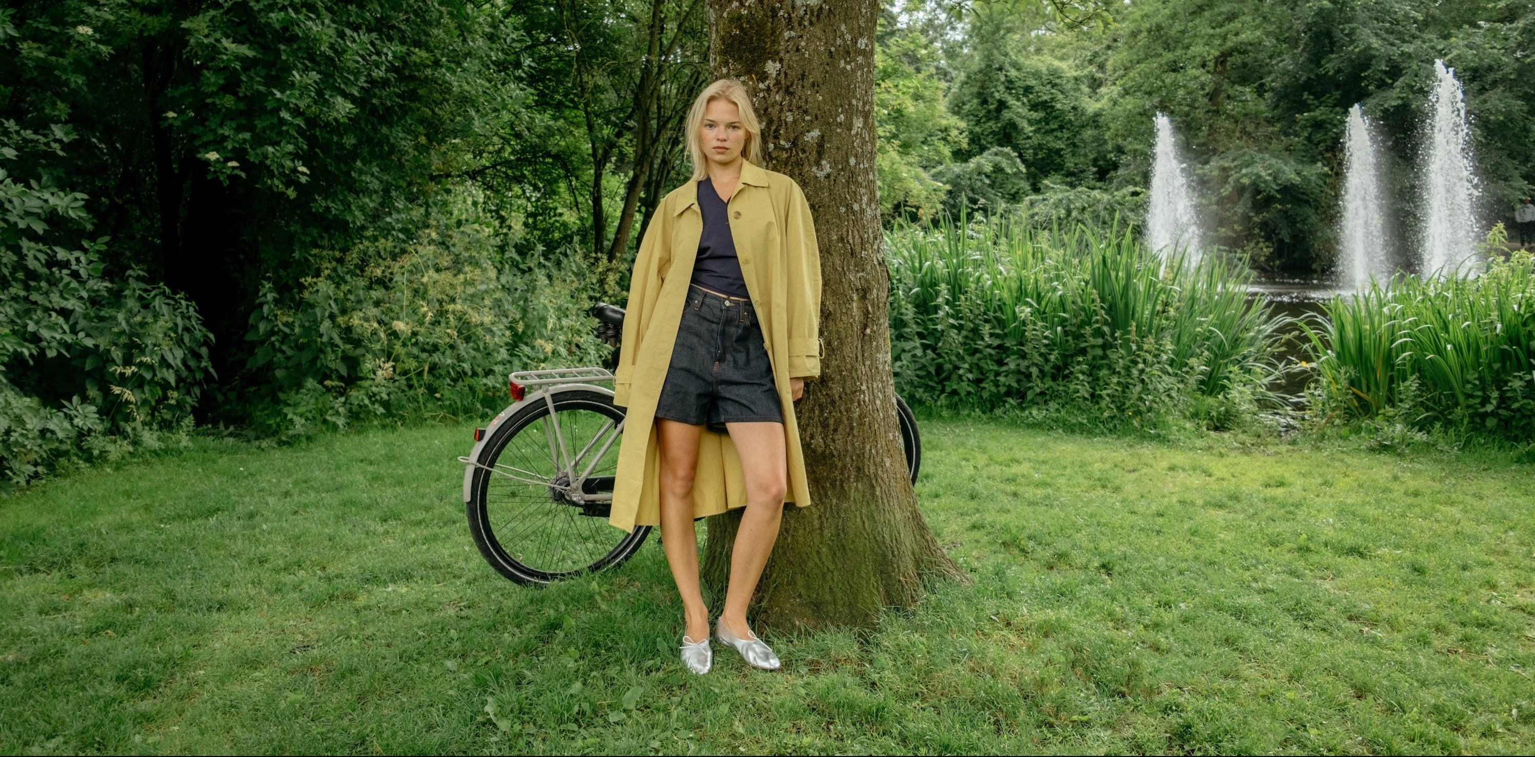 A woman stands outdoors in a park next to a large tree, wearing a long mustard yellow trench coat over a dark blue top and denim shorts. A bicycle is parked behind her, and a fountain is visible in the background among green trees and grass.