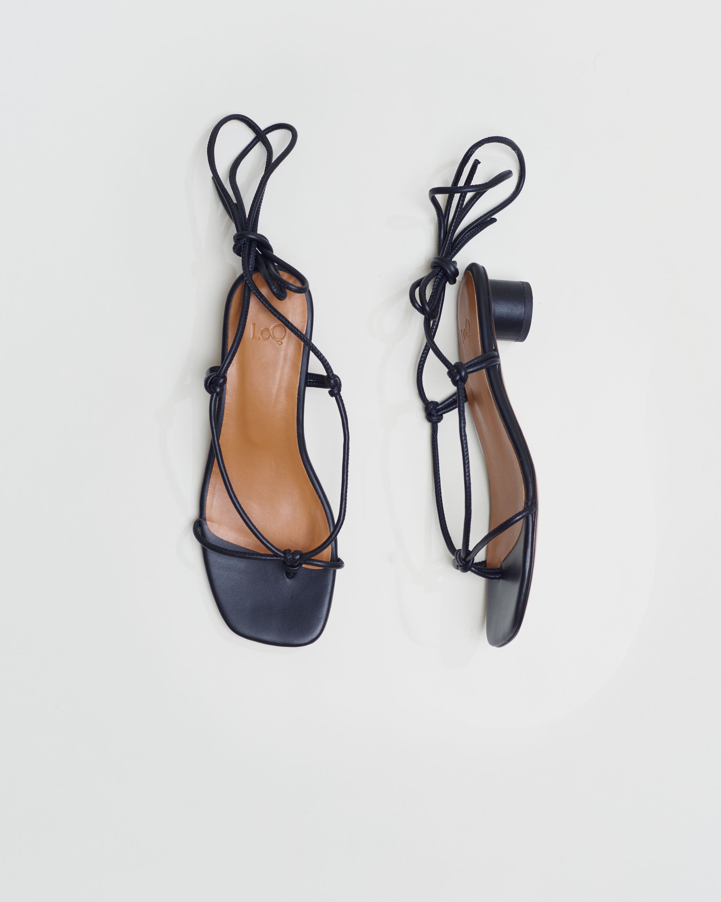 Knotted thong sandal featuring a short heel and tubular black leather straps that wrap around the ankle to tie.