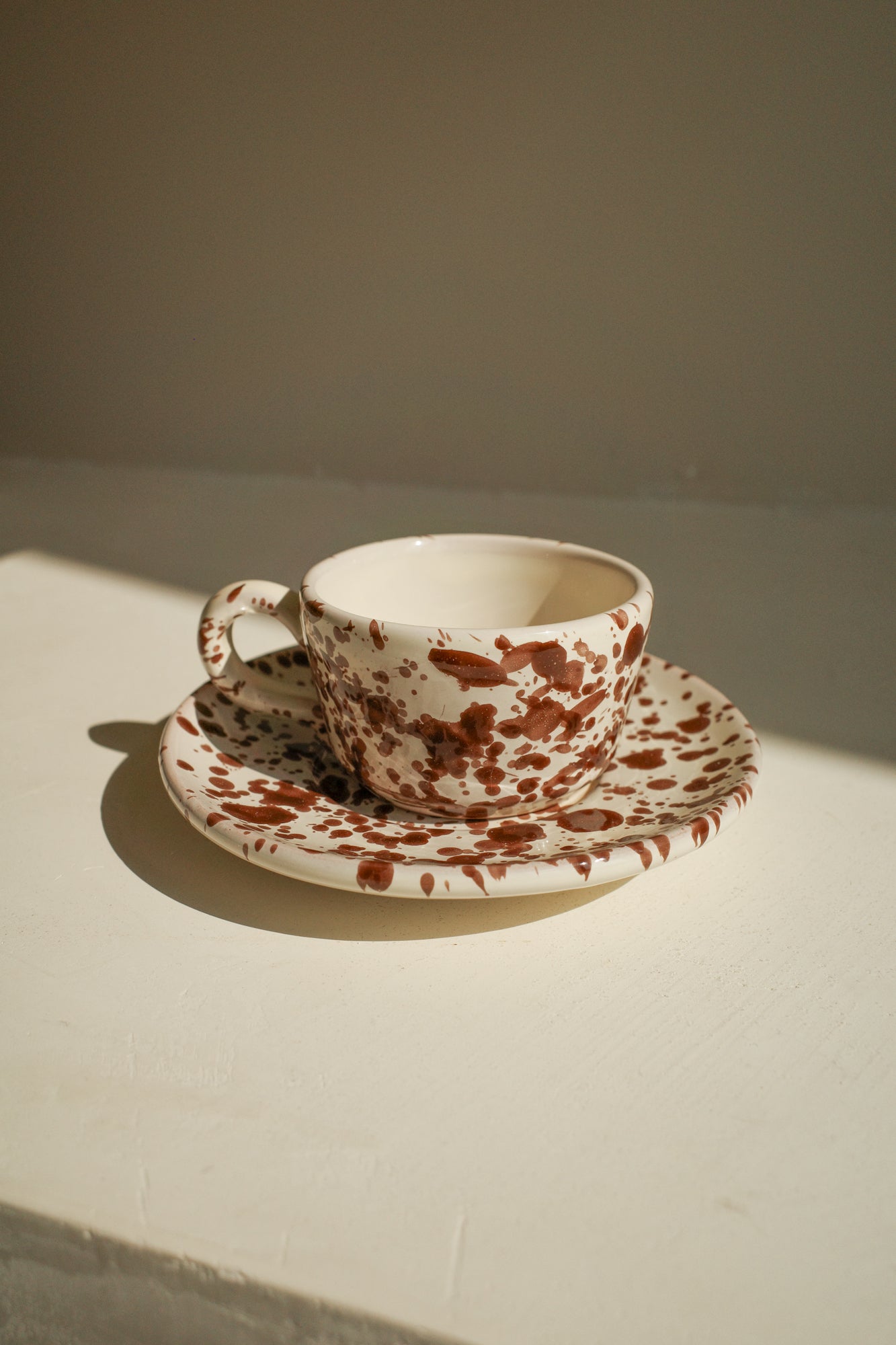 Ceramic espresso cupsand saucer with white glaze and dark brown splatter pattern.